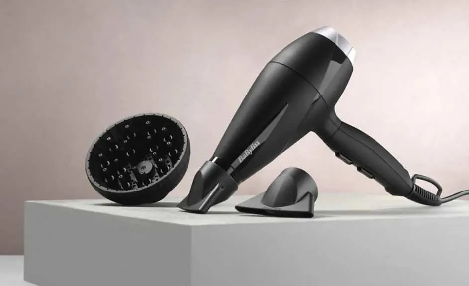 Un sèche-cheveux noir avec embouts sur une surface blanche, fond rose. Le sèche-cheveux est de marque Babyliss.
