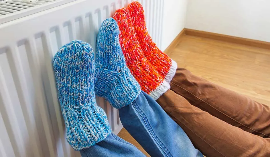 Twee paar voeten met gebreide sokken rusten tegen een witte radiator. Een paar is blauw, het andere oranje. Ze dragen jeans en bruine broeken.