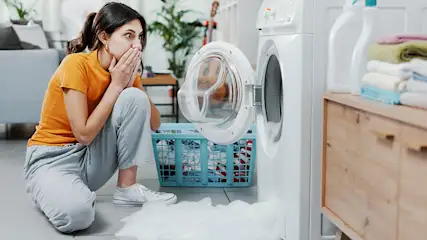 Een wasmachine met open deur en een blauwe wasmand. Er ligt schuim op de grond. Op een kast staan wasproducten en handdoeken.