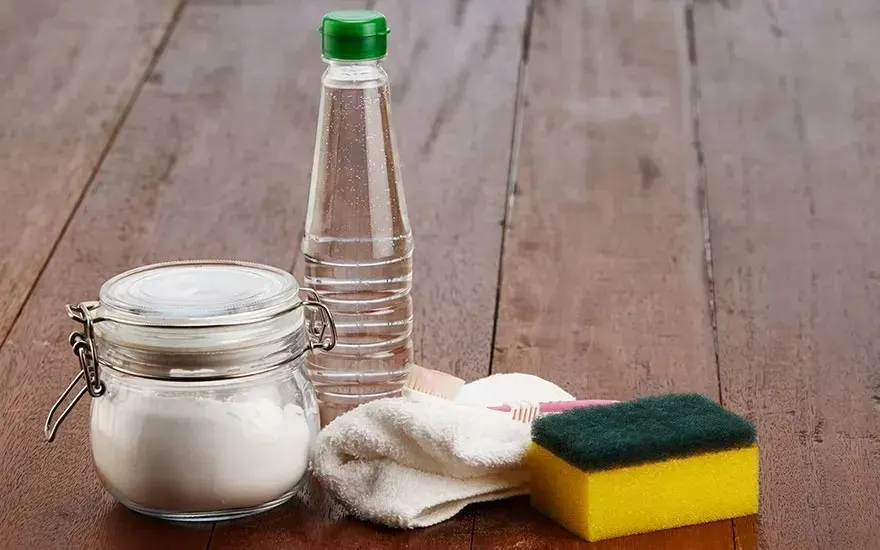Een glazen pot met wit poeder, een fles azijn, een spons, een doek en een tandenborstel liggen op een houten tafel, geen merk zichtbaar.