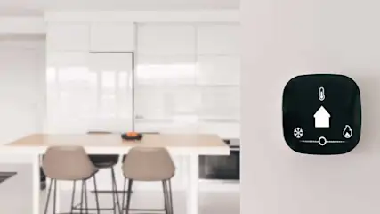 Zwarte thermostaat met witte symbolen (temperatuur, huis, sneeuwvlok, vlam) aan een witte muur. Achtergrond: keuken met witte kasten en een tafel.