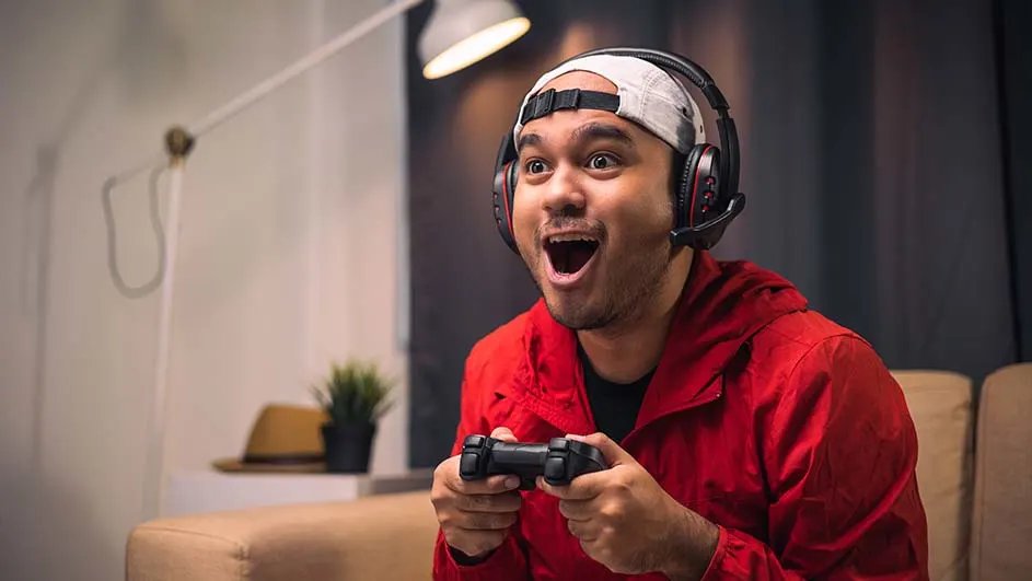 Un homme en veste rouge joue avec une manette noire. Un casque audio est sur sa tête. Une lampe est visible en arrière-plan.