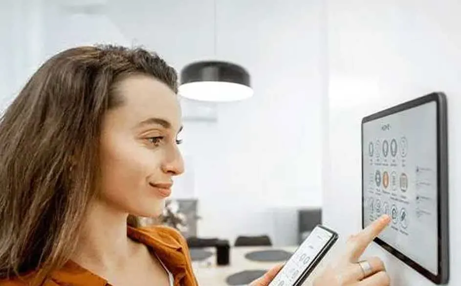 Een witte muur met een digitaal scherm toont diverse pictogrammen. Een hand tikt op het scherm, terwijl een smartphone in de andere hand wordt gehouden.