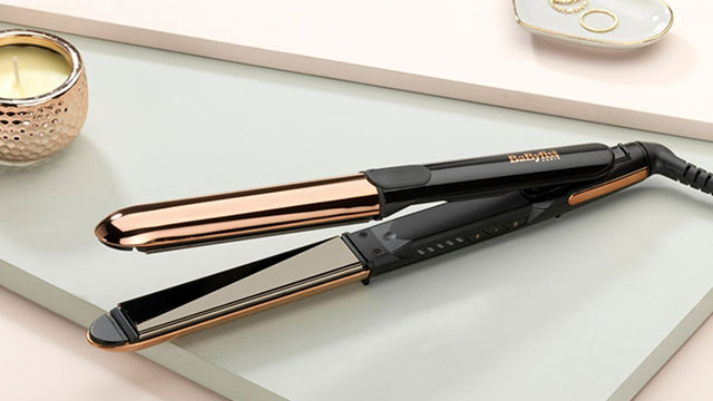 A black and bronze hair straightener sits on a white surface, near a candle and jewelry dish.