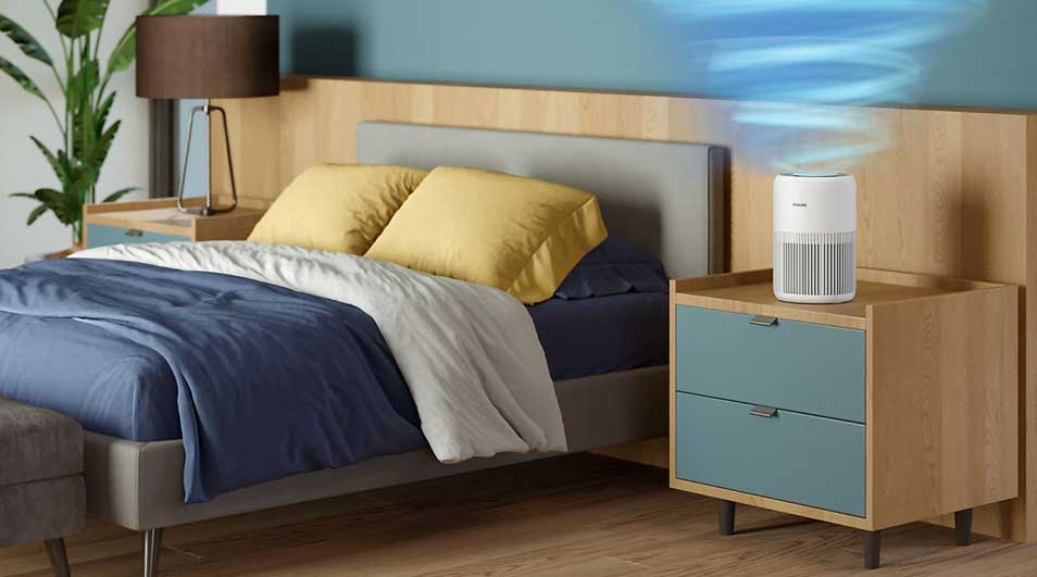 Slaapkamer met houten bed en nachtkastjes; een witte luchtreiniger blaast blauwe wervels boven het kussen in rustige, opgeruimde ruimte.