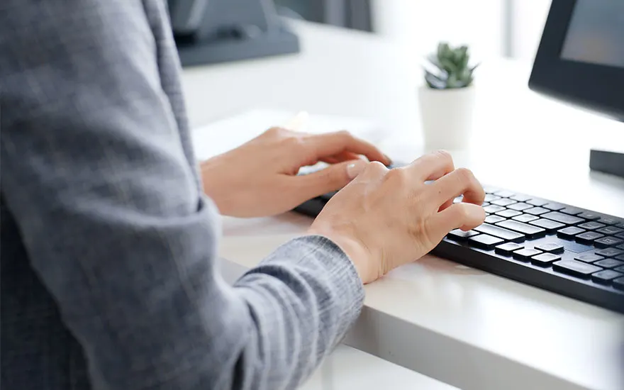 Mains tapant sur un clavier noir sur un bureau blanc. Un écran d'ordinateur et une plante sont visibles en arrière-plan.