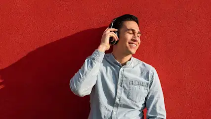Een man met een lichtblauw hemd luistert naar een hoofdtelefoon tegen een rode muur.