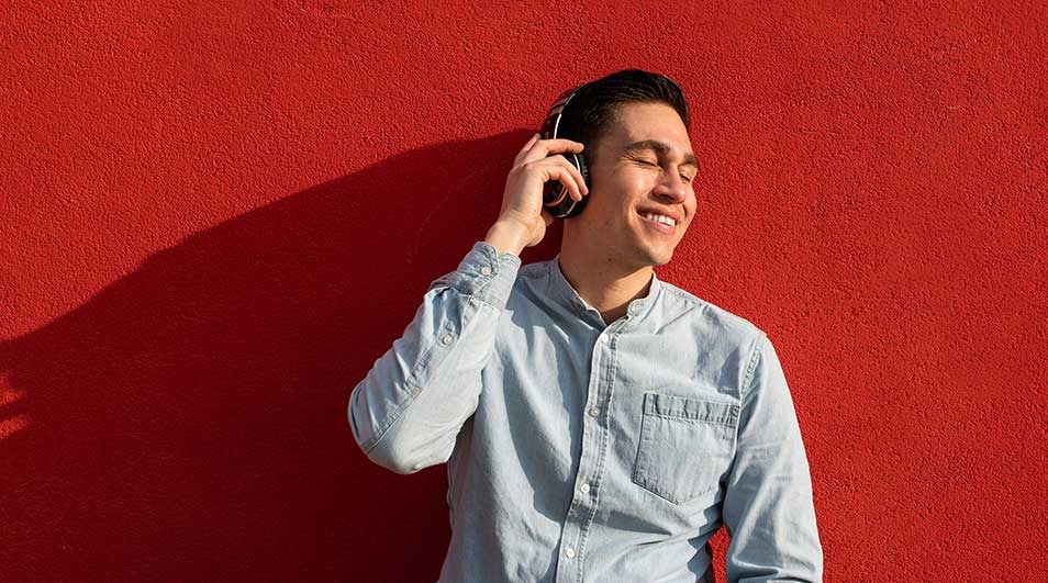 A person with headphones stands against a red wall, a shadow visible on the wall. The person wears a light blue shirt.