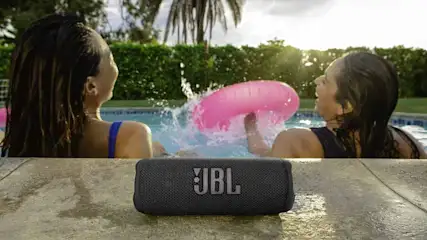 Haut-parleur JBL noir sur pierre au bord de la piscine. Bouée rose et éclaboussures d'eau en arrière-plan.