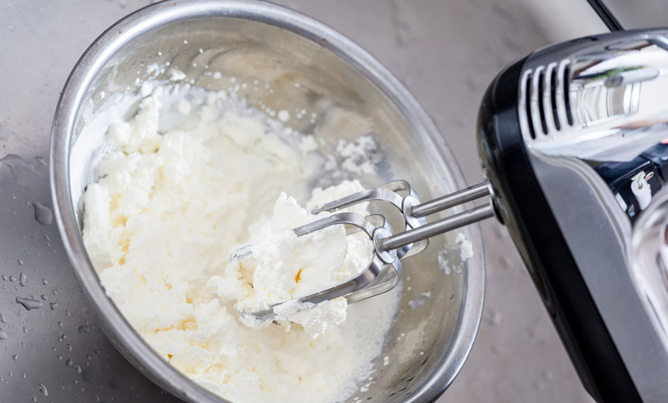 Een zilveren kom gevuld met witte substantie wordt gemengd door een elektrische mixer met metalen kloppers.