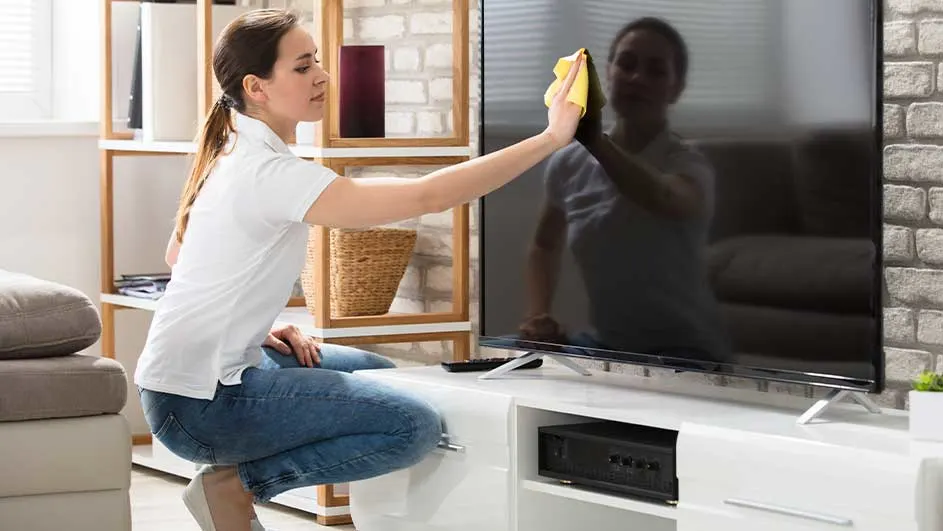 Une personne nettoie un grand téléviseur noir avec un chiffon jaune dans un salon. Une étagère en bois et un mur de briques sont visibles.