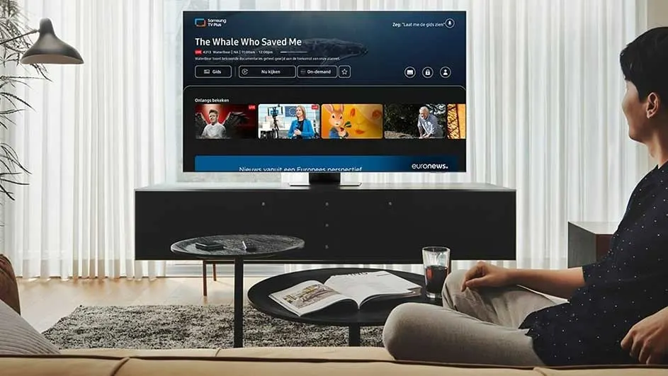 Woonkamer met tv die "The Whale Who Saved Me" toont, zwarte kast, salontafel met open boek en drankje, grijze vloerkleed en witte gordijnen.