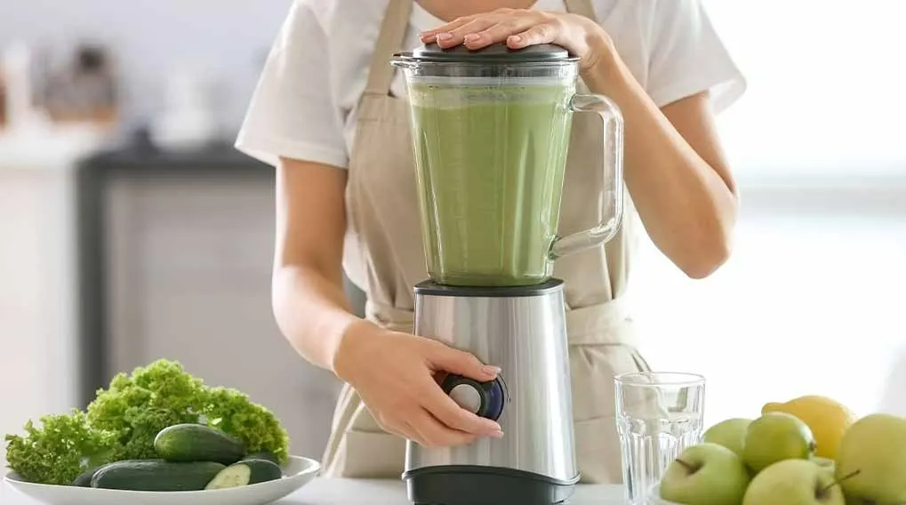 Une personne prépare un smoothie vert dans un mixeur en acier inoxydable, avec des fruits et légumes frais à proximité.