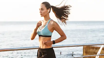 Een vrouw rent in sportkleding langs een reling, met de zee op de achtergrond. Ze draagt een sporthorloge en oortjes.