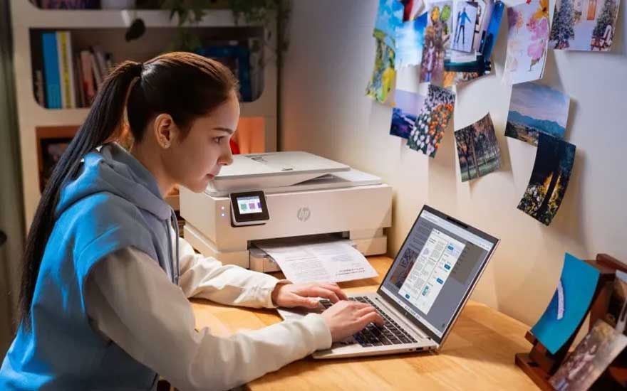 Een jonge vrouw print een document op haar bureau, terwijl een laptop openstaat. Foto’s en illustraties zijn als inspiratie aan de muur bevestigd.