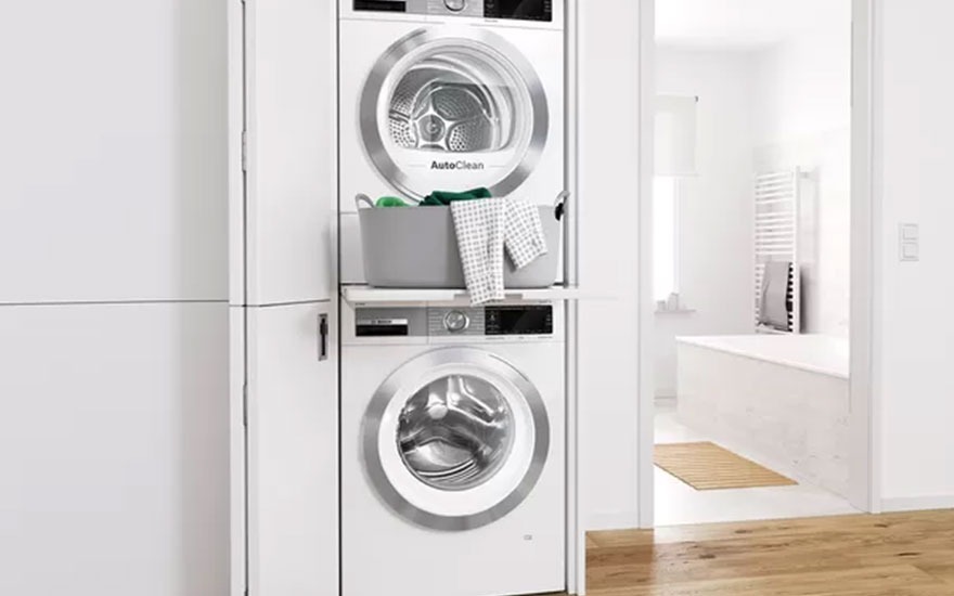 Stacked white washer and dryer unit next to a cabinet with a laundry basket. A bright bathroom is in the background.