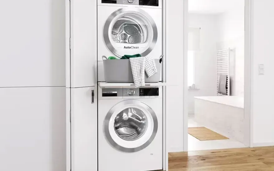 Stacked white washer and dryer unit next to a cabinet with a laundry basket. A bright bathroom is in the background.