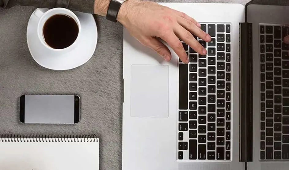 Main sur le clavier d’un MacBook Apple, à côté d’un café, d’un carnet et d’un iPhone ; design Apple bien identifiable.