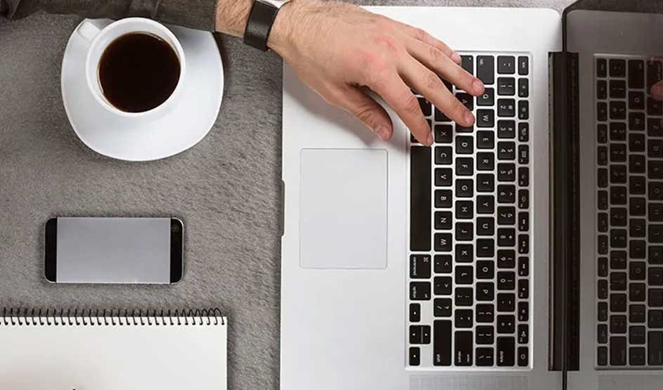 Top-down desk scene: a hand types on a silver laptop beside a cup of coffee, a smartphone, and a spiral notebook. No visible brand shown.