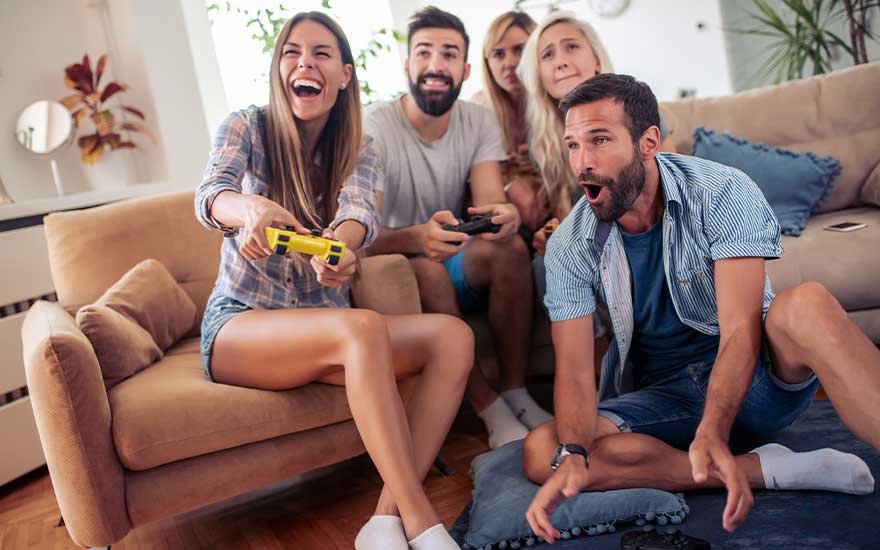 Cinq amis jouent à des jeux vidéo avec enthousiasme dans un salon confortable, riant et captivés.
