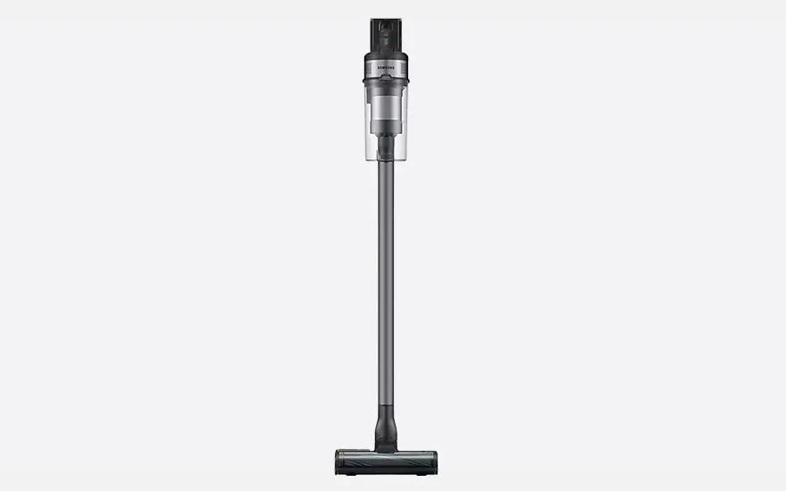 Slim grey stick vacuum stands upright against a light background, with clear dust container on top and a narrow floor nozzle at the bottom.