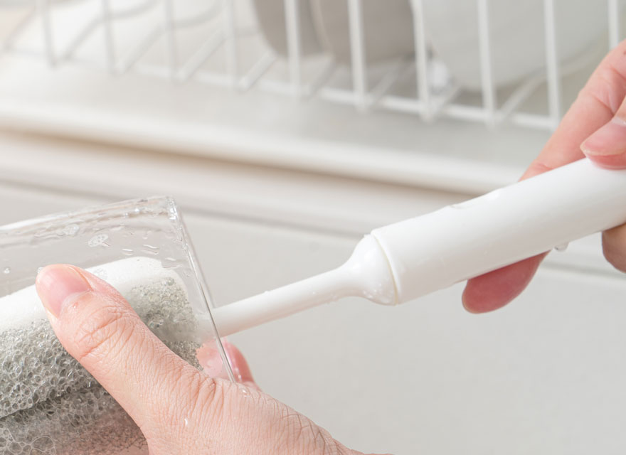 Une main tient une brosse blanche pour nettoyer un verre transparent sous l'eau, avec un égouttoir blanc en arrière-plan.