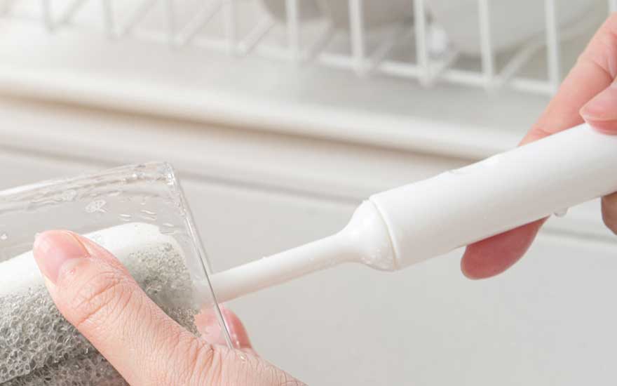 Close-up of hands washing a clear glass with a gray sponge and a white handled brush under running water, with a white rack behind.