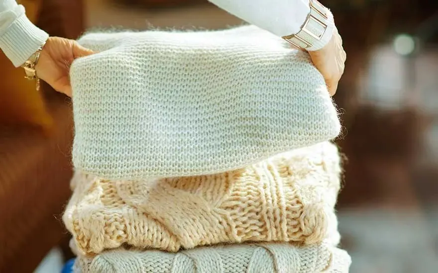 Een stapel beige gebreide truien wordt vastgehouden, met een witte trui bovenop.