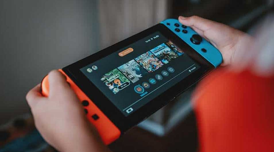 Hands hold a Nintendo Switch showing game icons on a black screen, with blue and orange Joy-Cons attached.