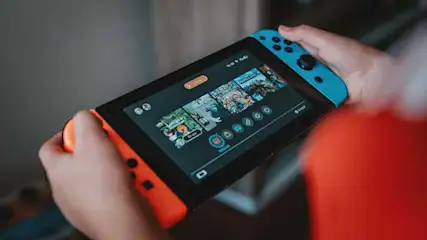 Hands hold a Nintendo Switch showing game icons on a black screen, with blue and orange Joy-Cons attached.