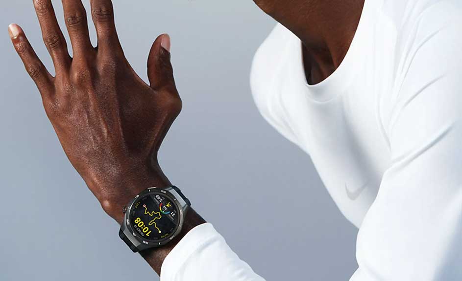 Un homme porte une montre noire affichant 10:08, avec un t-shirt blanc, sur un fond gris uni.