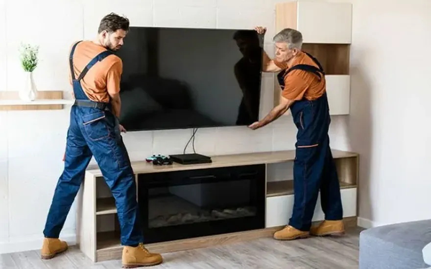 Twee mensen in blauwe overalls tillen een grote tv boven een tv-meubel met open haard.