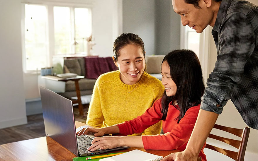 Een gezin zit aan een houten tafel met een laptop, notitieboek en potlood. Licht komt van een raam in de achtergrond.