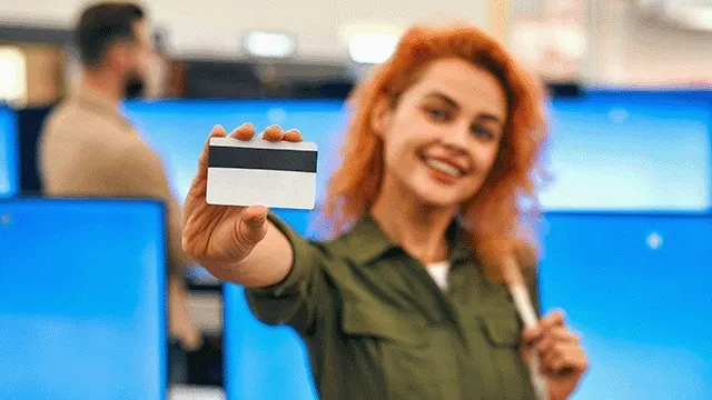 Een witte kaart met een zwarte magnetische strip wordt vooraan getoond, tegen een achtergrond van blauwe schermen in een winkel.