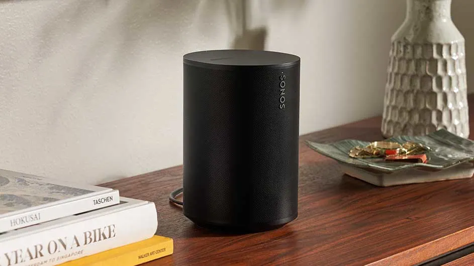 Een zwarte Sonos-luidspreker staat op een houten tafel, met boeken, een vaas en een schaaltje met sieraden op de achtergrond.