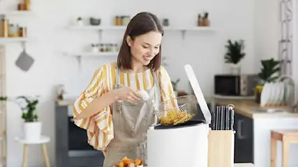 Une friteuse blanche contient un panier avec des frites. Des couteaux sont à côté, dans une cuisine claire.