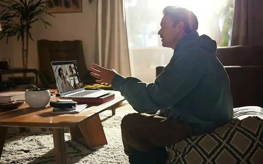 Man at home on cushion, video calling on laptop by low wooden table with coffee, sunlight filling the cozy room.