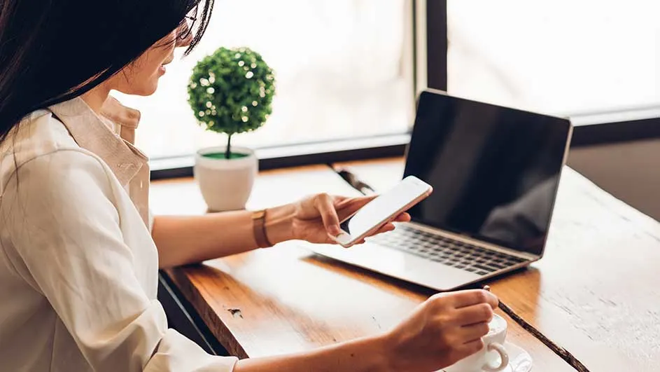 Een houten bureau met een laptop, een mobiele telefoon, een kopje en een groene plant in een witte pot.