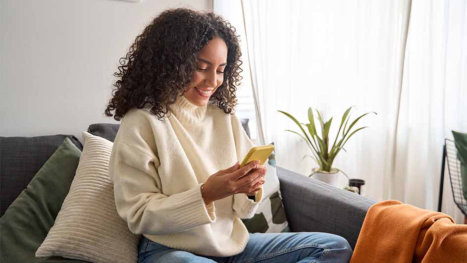 Une personne est assise sur un canapé gris avec des coussins, regardant un téléphone jaune. Une plante et une couverture orange sont visibles.