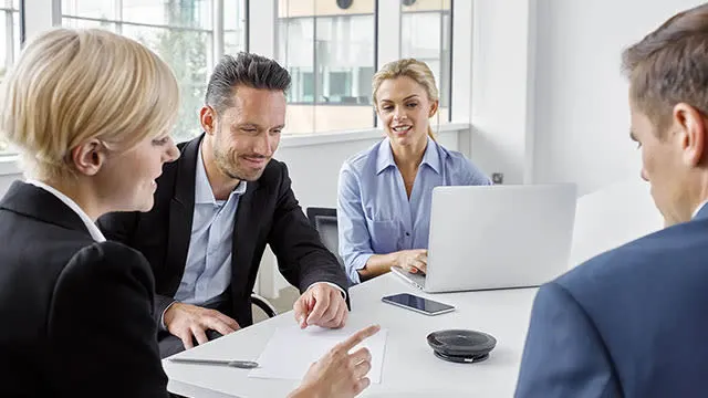 Quatre personnes sont assises autour d'une table blanche avec un ordinateur portable, un smartphone, un stylo et un Enceinte.