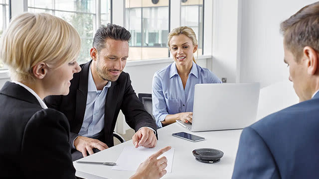 Een lichte tafel met een zwarte speakerphone, een smartphone, een pen en documenten. Een laptop staat open. Achtergrond met ramen.