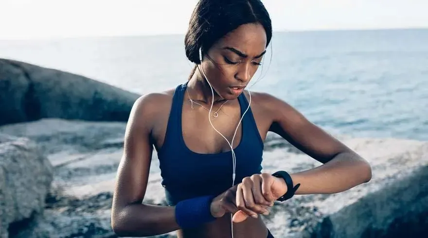 Une femme en tenue de sport bleue regarde sa montre connectée sur fond de mer et de rochers. Écouteurs blancs visibles.