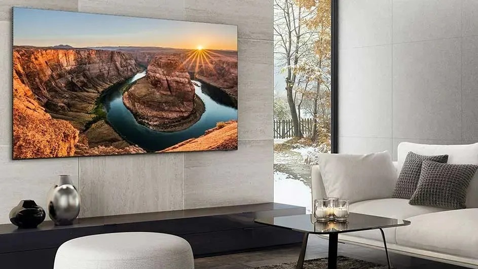 Modern living room with a big wall-mounted TV showing a canyon sunset, beside a tall window framing snowy trees.