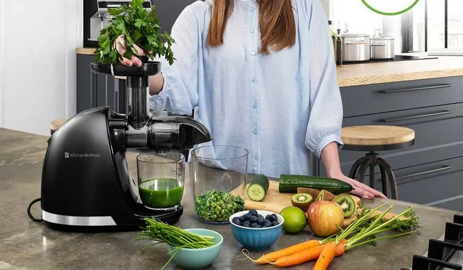 Une femme met du persil frais dans une centrifugeuse. Sur le plan de travail, il y a concombre, carottes, pommes, kiwi, citron vert et myrtilles.