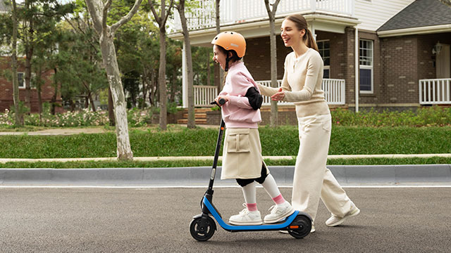Une trottinette bleue sur une route goudronnée, avec une bordure grise, de l'herbe verte et des arbres en arrière-plan.