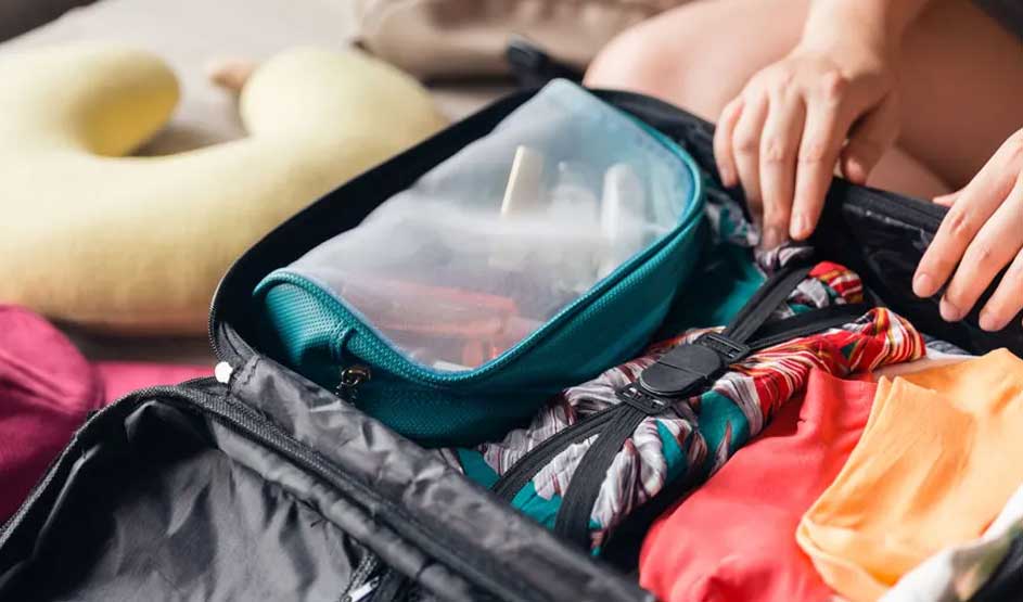 Valise noire ouverte remplie de vêtements colorés et d'une trousse de toilette turquoise. Oreiller de voyage jaune à côté.