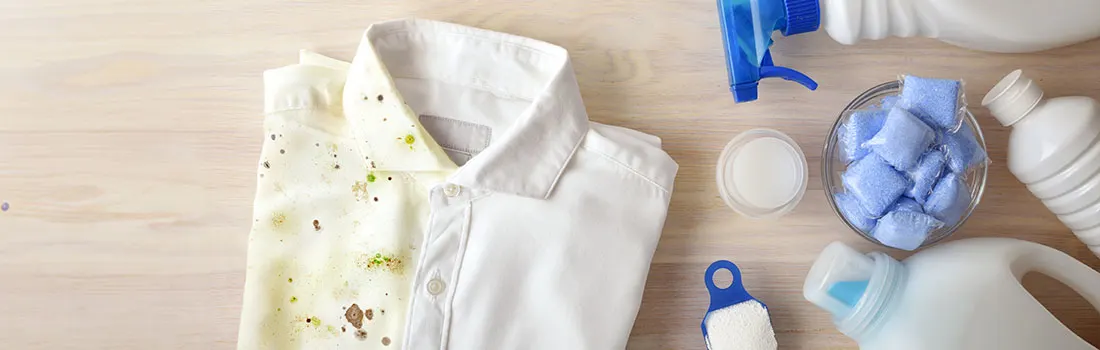 Vue de dessus : chemise tachée à côté de détergent, spray et capsules bleues sur bois clair.
