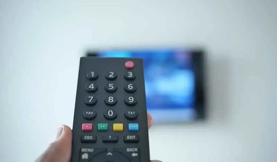 Een hand houdt een zwarte afstandsbediening gericht op een onscherpe tv op de achtergrond, klaar om een kanaal te wisselen.