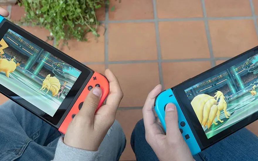 Twee mensen zitten buiten en spelen samen Pokémon op hun Nintendo Switch-consoles, waarbij ze elk kleurrijke controllers vasthouden.