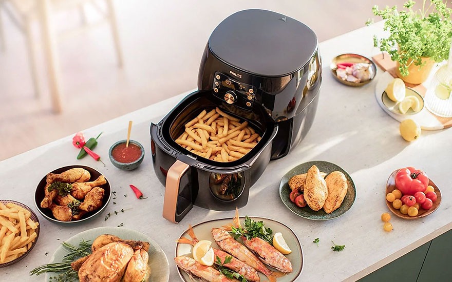 Une friteuse noire avec des frites, entourée de plats de poulet, poisson, tomates et herbes sur une table blanche.